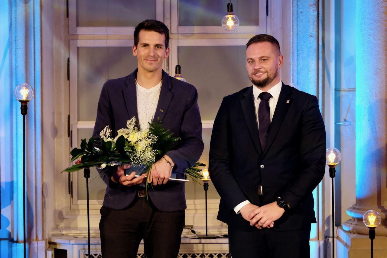dvaja slušne oblečení muži stoja, muž vľavo má v rukách kyticu a diplom, muž v pravo nemá nič